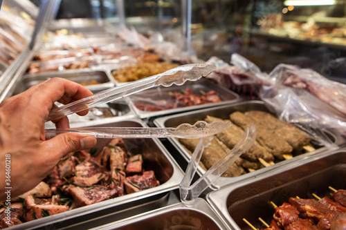 Selective focus view of butcher hand holding plastic tong with a variety of meat products ready for cooking on the display refrigerator in background.