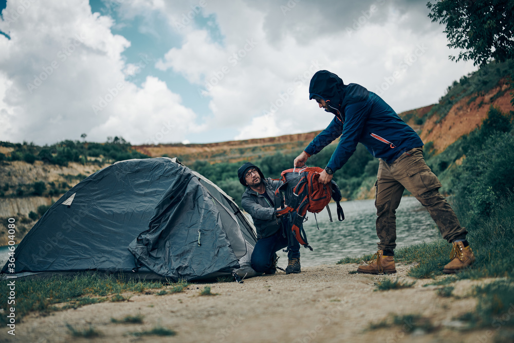Fototapeta premium Two men on camping trip putting backpacks into tent by the lake on rainy and cold weather.