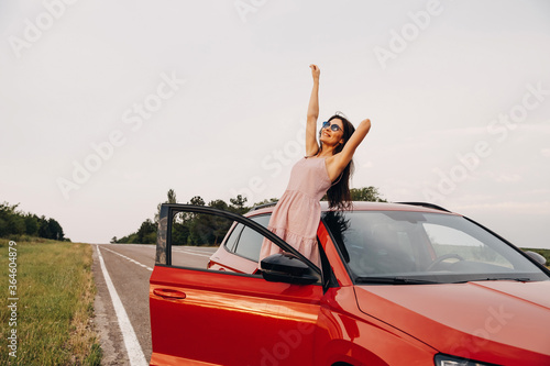 Young beautiful woman with hands up in the air, enjoying summer wind next to her car. Summer vacation concept.