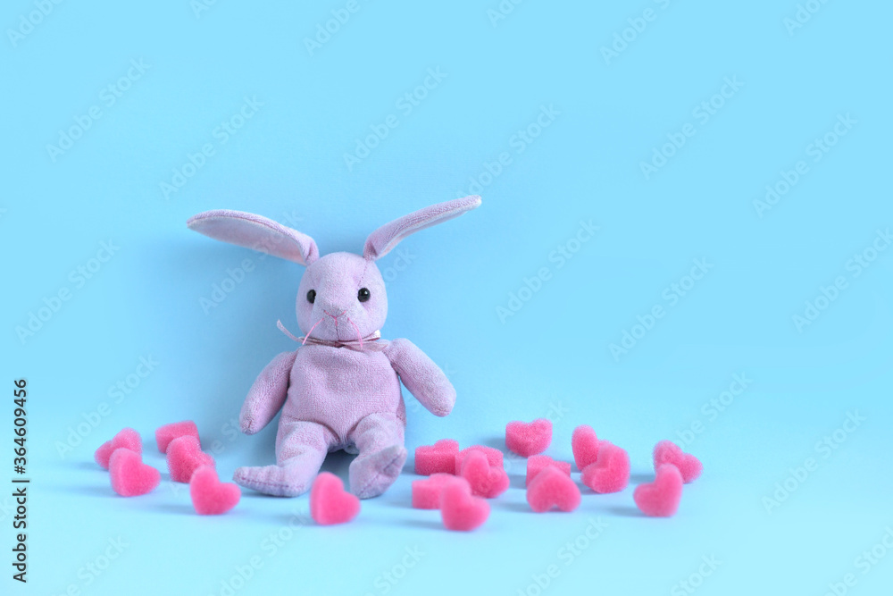 Obraz premium Pink plush hare sits on blue background among soft pink hearts. Valentine's day card