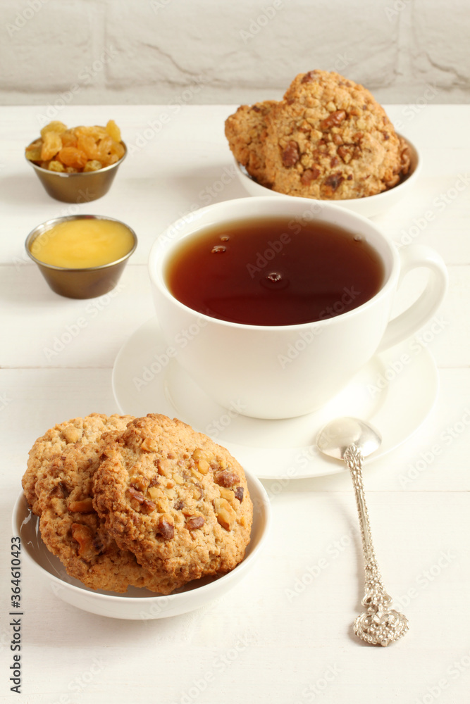 Oatmeal cookies with walnuts and raisins on small saucers and a cup of black tea, standing on a white wooden table against a white brick wall. Closeup Oatmeal cookies with walnuts and raisins on small saucers and a cup of black tea, standing on a white wooden table against a white brick wall. Closeup