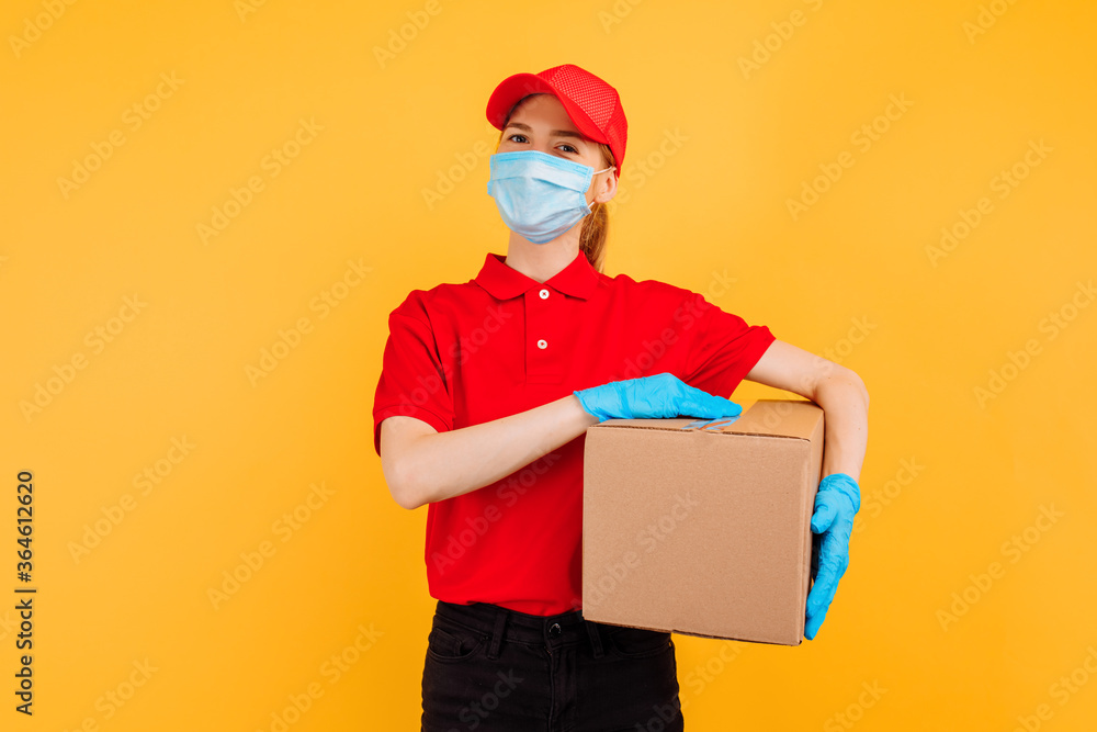 young delivery girl in a red uniform, wearing a medical mask and gloves, holds a cardboard box isolated on a yellow background. Delivery service, quarantine, coronavirus young delivery girl in a red uniform, wearing a medical mask and gloves, holds a cardboard box isolated on a yellow background. Delivery service, quarantine, coronavirus