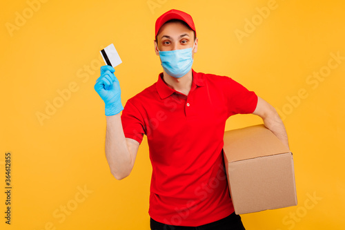 Courier-a man in a medical mask and gloves, in a red cap, t-shirt, uniform. A delivery man holds a cardboard box and shows a credit card. Online stores, delivery service