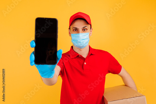 male courier in a red uniform, medical mask and gloves holds a cardboard box and shows an empty mobile phone screen on a yellow background. Delivery service, online stores, coronavirus