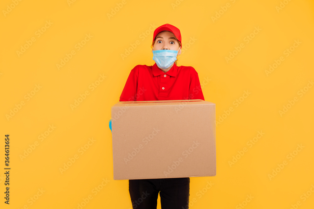 shocked surprised delivery woman wearing a medical mask on her face, holding and carrying a cardboard box isolated on a yellow background. Delivery service, quarantine shocked surprised delivery woman wearing a medical mask on her face, holding and carrying a cardboard box isolated on a yellow background. Delivery service, quarantine