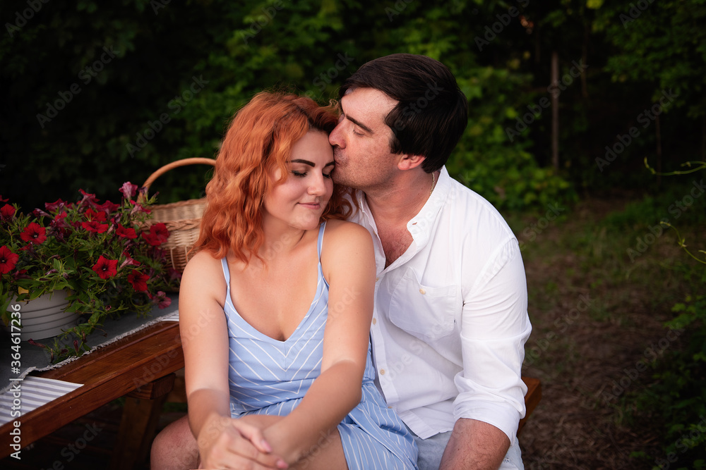 Close-up portrait of a couple in love, resting on a picnic at a wooden table with a basket of food. A young brunette man gently kisses a beautiful red-haired girl on the cheek, she laughs. Travel love Close-up portrait of a couple in love, resting on a picnic at a wooden table with a basket of food. A young brunette man gently kisses a beautiful red-haired girl on the cheek, she laughs. Travel love