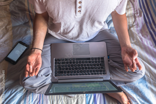 Bird's eye angle photography of a homeoffice with a young barefoot man in pajamas and meditation pose studying/working with a laptop on his legs. Blanket with blue and purple stripes background. photo