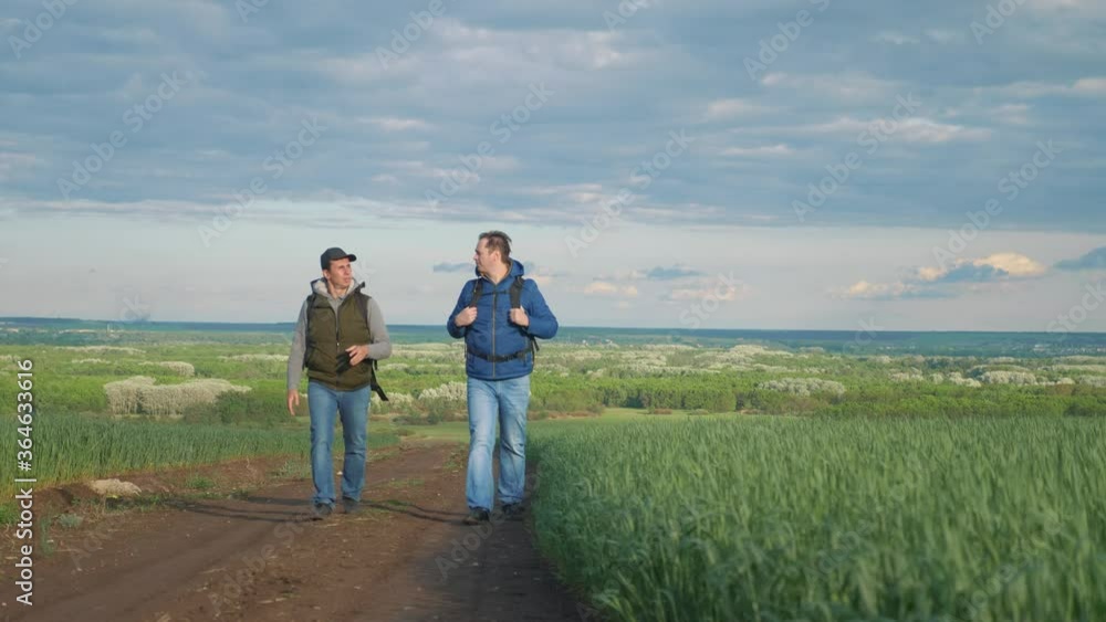 teamwork travel. two man hikers with backpacks walk lifestyle along trail next to a field with green grass in nature. concept travel adventure hiking healthy. two tourists walk lifestyle walking teamwork travel. two man hikers with backpacks walk lifestyle along trail next to a field with green grass in nature. concept travel adventure hiking healthy. two tourists walk lifestyle walking