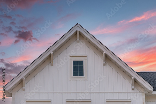 New gable with decorative brackets, white vertical vinyl lap siding with square attic window on a newly built home in America colorful sunset background 