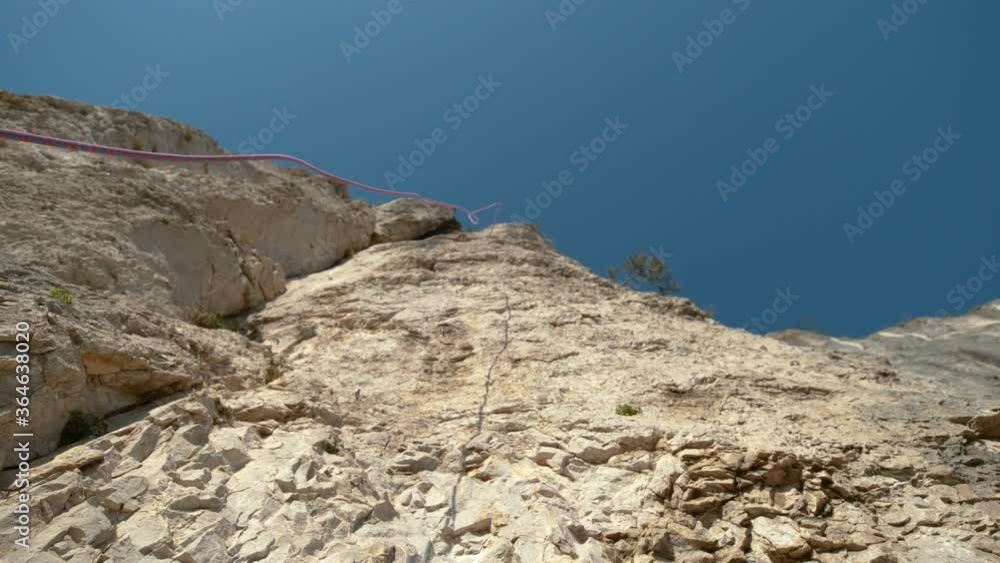 SLOW MOTION, BOTTOM UP, DOF: Blue and red rope falls from the top of a towering cliff on a sunny summer day. Safety rope gets released after a group of rock climbers all reach the top of a rocky wall. SLOW MOTION, BOTTOM UP, DOF: Blue and red rope falls from the top of a towering cliff on a sunny summer day. Safety rope gets released after a group of rock climbers all reach the top of a rocky wall.