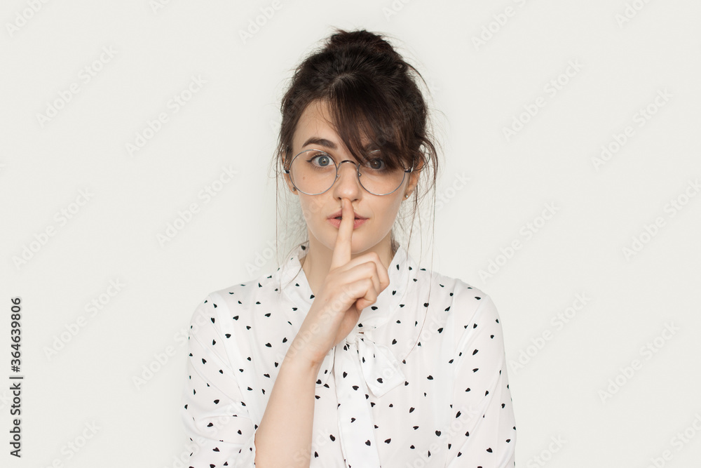 Lovely brunette lady with eyeglasses gesturing the silence sign on a white studio wall looking at camera Lovely brunette lady with eyeglasses gesturing the silence sign on a white studio wall looking at camera
