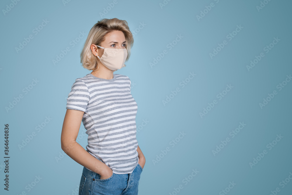 Fashionable blonde woman with medical mask on face posing on blue studio wall with free space Fashionable blonde woman with medical mask on face posing on blue studio wall with free space