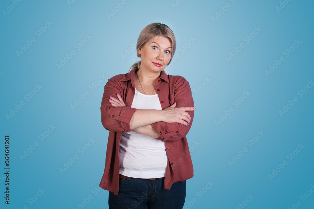Interested caucasian senior woman with blonde hair posing with crossed hands on blue studio wall looking at camera Interested caucasian senior woman with blonde hair posing with crossed hands on blue studio wall looking at camera