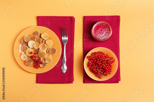 Sweet mini pancakes in an orange plate stand on a red napkin, next to it is a jar of red currant jam and a cup of red currant, orange background, flat lay, breakfast concept, knolling..