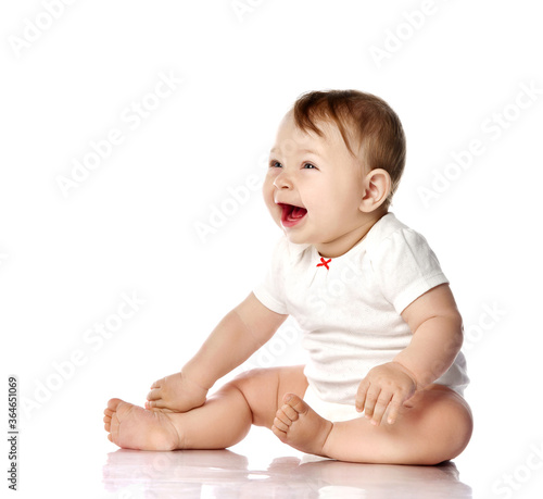 Laughing loud adorable little baby infant toddler in white cotton bodysuit sits on floor touching legs, looking aside
