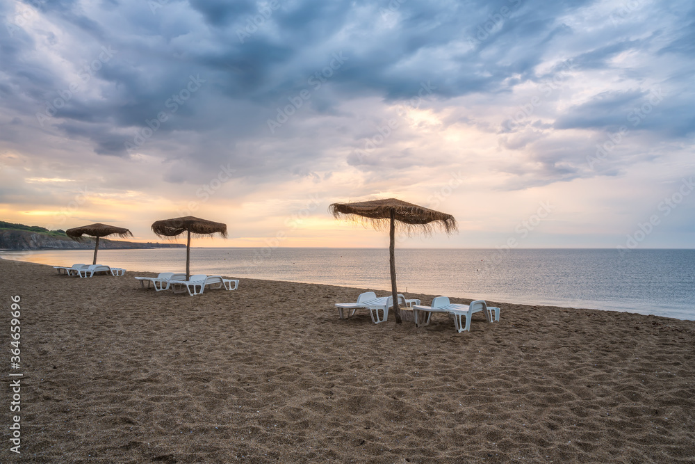 Straw sunshades and sunbeds on the empty sandy beach with sea in the background before sunset Straw sunshades and sunbeds on the empty sandy beach with sea in the background before sunset