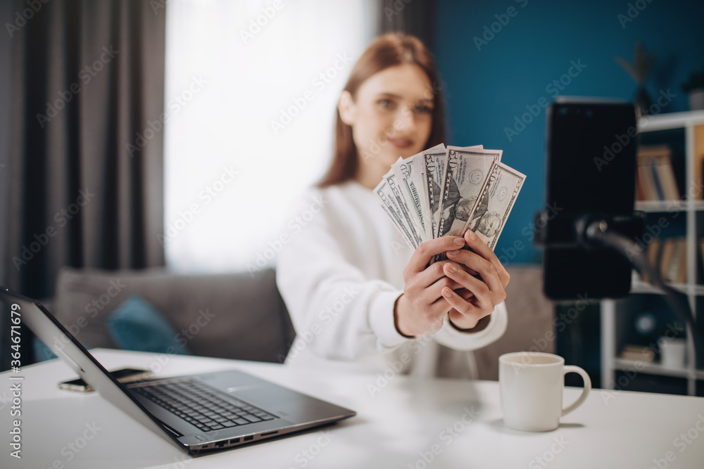 Successful blogger with brown hair showing on camera dollars banknotes while sitting at table. Attractive girl explaining her followers in social networks how to be satisfied with salary. Successful blogger with brown hair showing on camera dollars banknotes while sitting at table. Attractive girl explaining her followers in social networks how to be satisfied with salary.