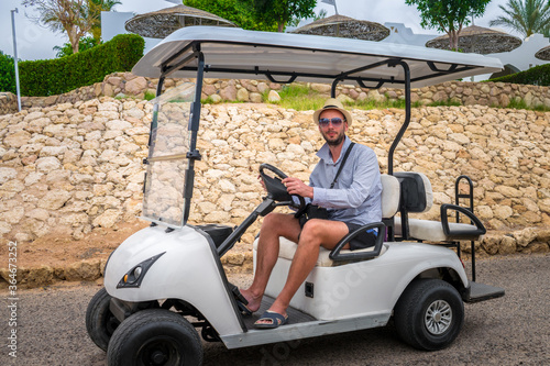 A guy in a hat and glasses rides a golf cart on an Arab street. A man sits behind the wheel of an electric car in a tropical resort.