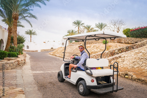A man sits behind the wheel of an electric car in a tropical resort. A guy in a hat and glasses rides a golf cart on an Arab street