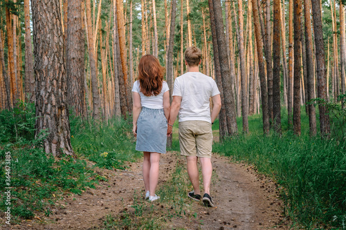 A blond man and a girl with long hair in a skirt walk hand in hand through a pine forest