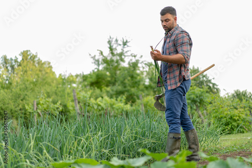 Young man with hoe in his garden. Holding and looking spring onion from his organic farm. Concept eco farm vegetable garden.