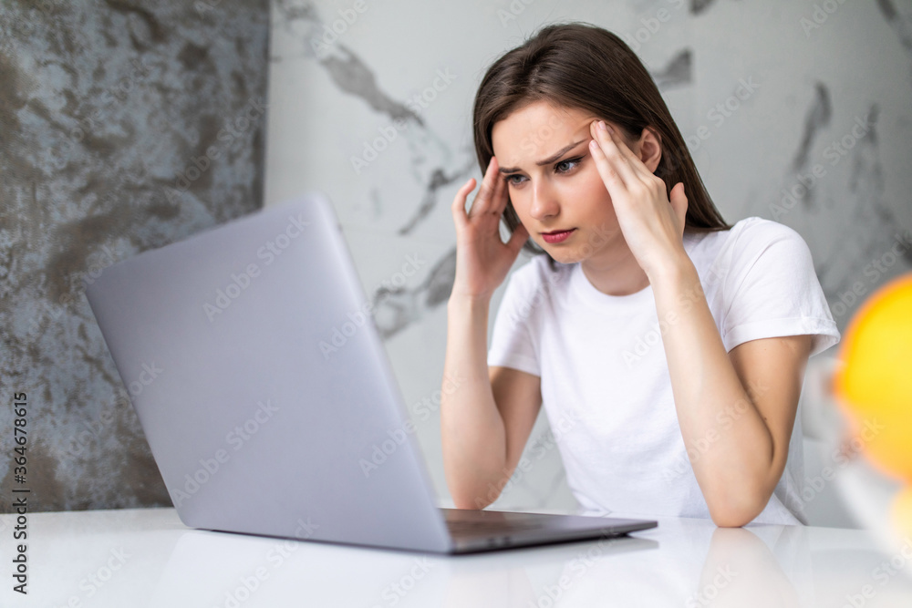 Young pretty woman having headache while working on laptop sitting at her kitchen table at home. Young pretty woman having headache while working on laptop sitting at her kitchen table at home.