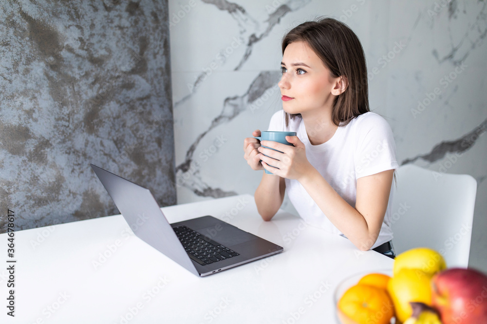 Smiling young woman with coffee using laptop in the kitchen at home. Smiling young woman with coffee using laptop in the kitchen at home.
