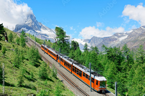 Summer scenery of a cogwheel train of Gornergrat railway traveling thru a forest on the hillside with majestic Matterhorn mountain peak veiled by clouds under blue sky, in Zermatt, Valais, Switzerland