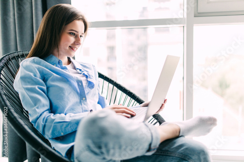 Young smiling brunette girl is sitting on modern chair near the window in light cozy room at home working on laptop in relaxing atmosphere