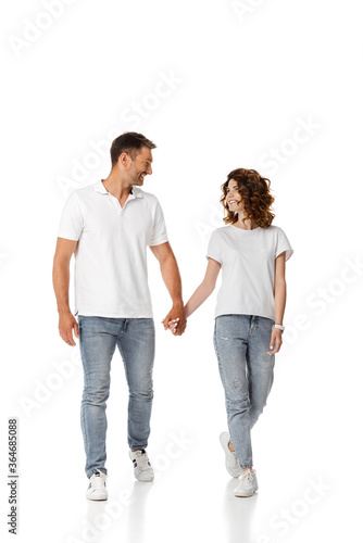 happy woman and cheerful man holding hands and looking at each other while walking on white