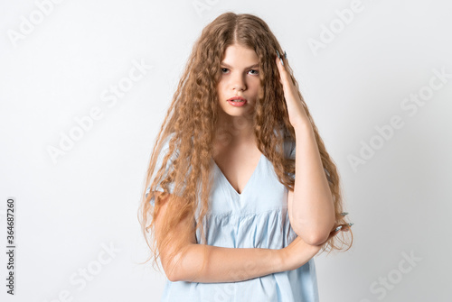 Photo of tired European young woman with curly long hair, holds his hand near his head, feels fatigue, needs good rest, has sleepy expression, isolated over white background. Weariness and people