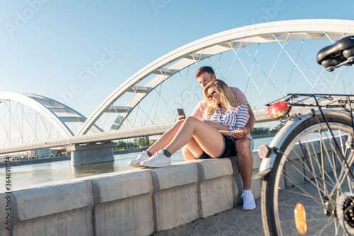 Young couple with bicycle enjoying together sitting by the river and embracing each other and making selfie. © Zoran Zeremski