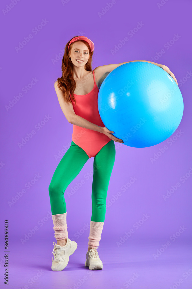sporty student girl holding blue fitness ball standing relaxed against purple background, looking stylish in cute pink bodysuit, green leggins, sun cap and white sport shoes. Full lenght view sporty student girl holding blue fitness ball standing relaxed against purple background, looking stylish in cute pink bodysuit, green leggins, sun cap and white sport shoes. Full lenght view