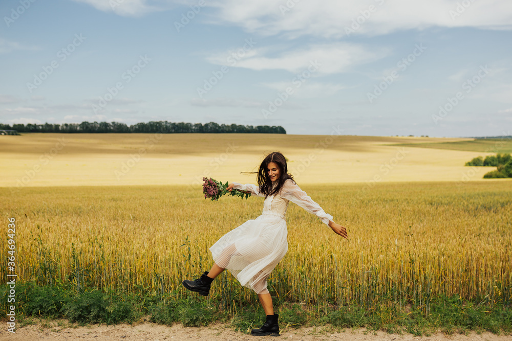 Woman in beautiful white dress jump in summer field with bouquet of flowers in hand. Long hair, tanned skin, emotions of happiness. Place for text.
