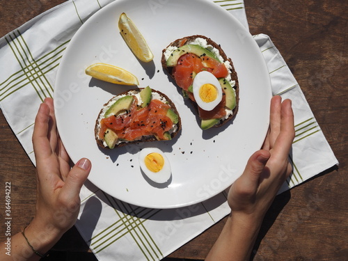 Healthy breakfast.Hands of a young woman holding a white plate with whole grain bread sandwiches, with cream cheese, egg, avocado and selmon fish. photo