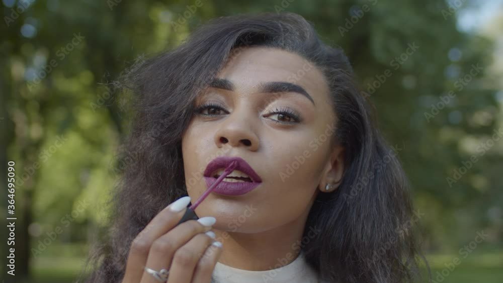 Lifestyle portrait of gorgeous charming african american woman with curly hair doing makeup, applying and correcting lipstick lip gloss while looking at camera as pocket mirror outdoors.