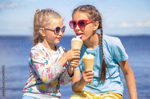 Two adorable little girls wearing sunglasses are eating ice cream on the seashore on summer vacation. Small girl is sharing her ice cream with sister