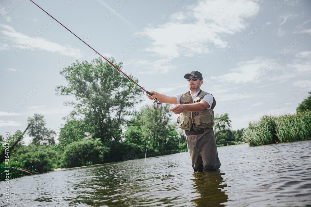 Young fisherman fishing on lake or river. Low view of guy in rob holding long rod and using it for catching fish. Stand in river or lake water alone. Fish hunting. Professional fisherman in action. Young fisherman fishing on lake or river. Low view of guy in rob holding long rod and using it for catching fish. Stand in river or lake water alone. Fish hunting. Professional fisherman in action.