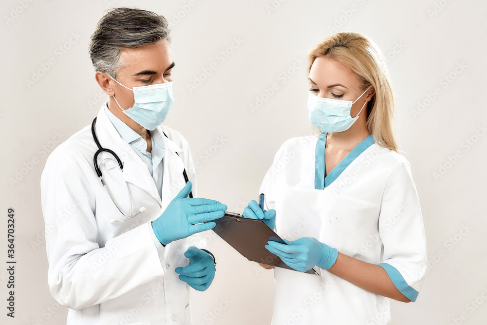 Two male and female doctors wearing medical uniforms and protective masks discussing laboratory test, standing against grey background Two male and female doctors wearing medical uniforms and protective masks discussing laboratory test, standing against grey background