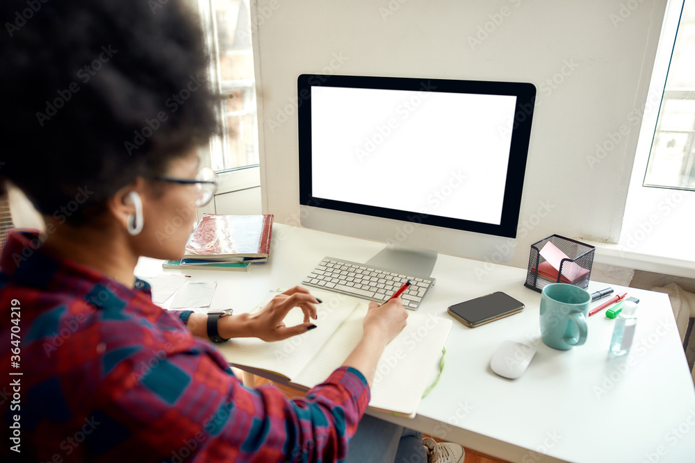 Rear view of afro american female student sitting at her workplace at home and studying online. Looking at blank computer screen and making notes