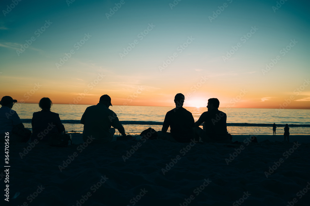 Young friends recreationg together on sandy beach in evening dusk enjoying vacation in summer, peoples silhouette looking at beautiful sunset and golden skyline over calm ocean water surface.