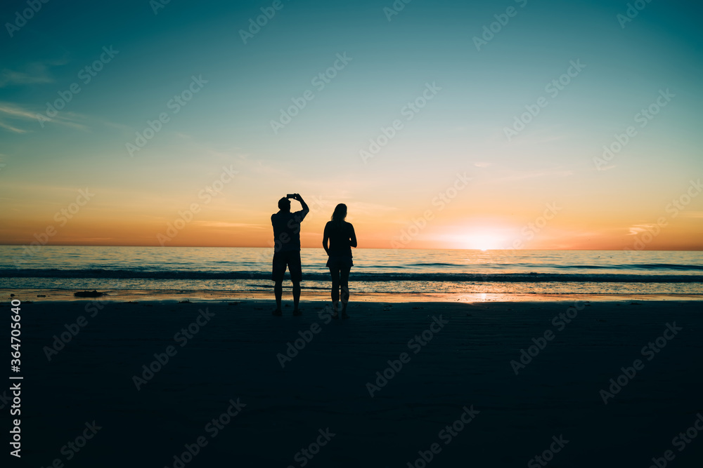 Male and female silhouettes on sandy shore of calm ocean during scenic sunset, couple in love enjoying beauty of evening tropical island view spending time on coast of sea during summer vacations.