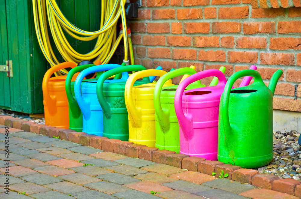 Many colourful watering cans standing in a line in front of a wall of red bricks Many colourful watering cans standing in a line in front of a wall of red bricks