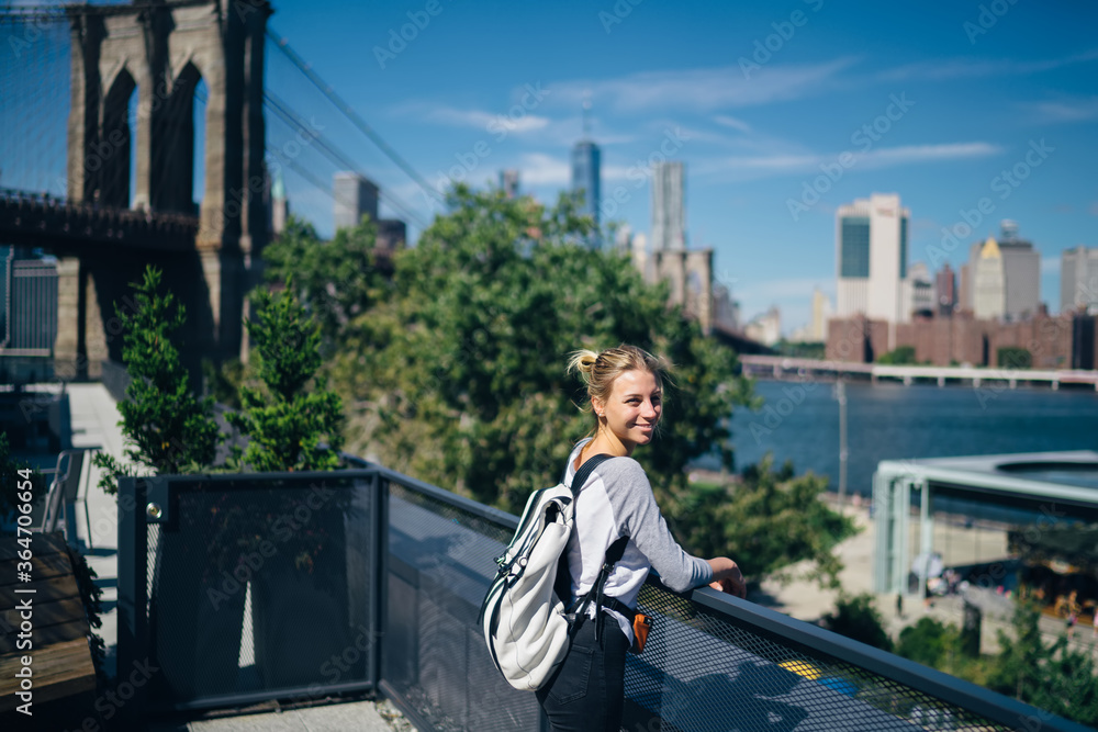 Obraz premium Positive young woman enjoying beautiful urban scenery near bridge over river during trip to America,smiling hipster girl with backpack having sightseeing tour of famous places in New York city.
