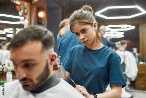 Focused professional barber girl working with hair clipper, making modern haircut for a young handsome man visiting barbershop