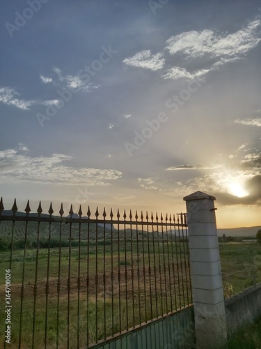 Fence lining with spikes. A photograph of ornate iron spikes on top of an old wall at sunset. Sunset behind blurry fence with cloudy red sky.