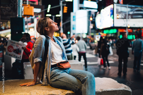Young female traveler fascinated with Time square lights sitting on crowded street, thoughtful hipster girl looking at modern architecture of famous New York landmark shining with neon commercial.
