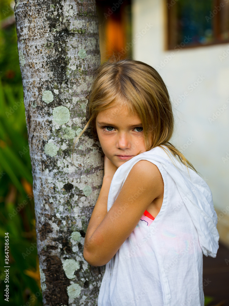 Gorgeous little girl standing near the tree, wearing white dress. Smiling face with pensive emotion. Spending time outdoor in tropical garden. Childhood concept. Gorgeous little girl standing near the tree, wearing white dress. Smiling face with pensive emotion. Spending time outdoor in tropical garden. Childhood concept.