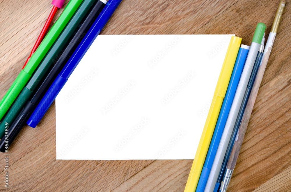 Obraz premium A white sheet of paper on a wooden background and stationery are nearby. Sheet of paper and pencils, felt-tip pens. Mock up