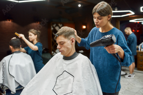 Working at barbershop. Professional young barber girl drying wet hair of her client sitting in in armchair in the modern barbershop